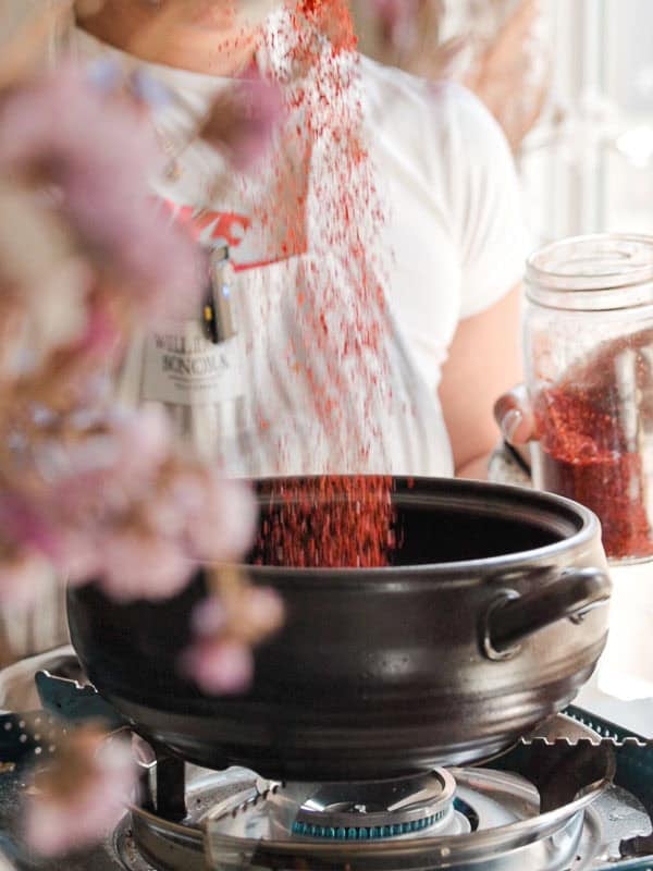 Person pouring gochugaru from a jar into a black pot on a gas stove. The pot has handles, and the person is wearing a white shirt. Blurred foreground elements include pinkish flowers.